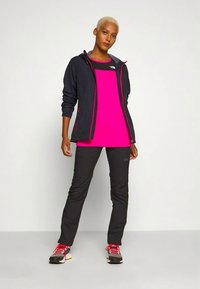 Kvinne med kort blondt hår som har på seg en svart jakke over en lys rosa topp, svarte bukser og grå joggesko med røde lisser, som står mot en hvit bakgrunn.