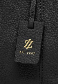 Etichetta in pelle nera testurizzata con logo dorato e "EST. 2007" inciso. Attaccata con una cinghia nera abbinata.