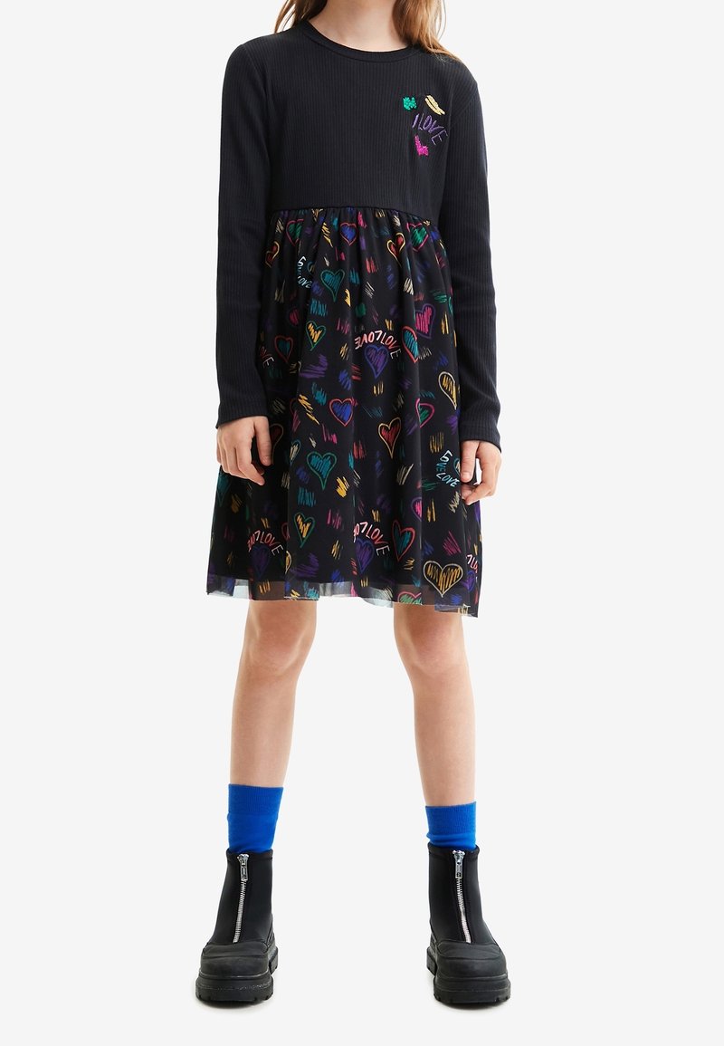 Fille portant une robe noire à manches longues avec des motifs de cœurs colorés et le texte « LOVE », associée à des chaussettes bleu vif et des bottes noires à fermeture éclair.