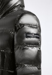 Zwarte pufferjas met een glanzende afwerking, voorzien van horizontale stiksels en een genaaid label op de linker schouder met de merknaam.