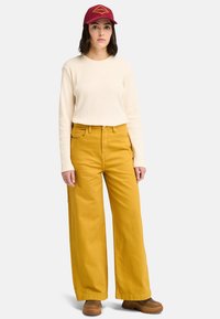 Krämfärgad långärmad ribbad topp, gula vida byxor, bruna skor och röd keps med broderad logotyp. En enfärgad bakgrund framhäver fokus på outfiten.