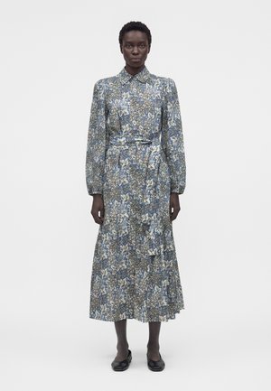 Žena stojí čelom vpred, oblečená v dlhých rukávoch, šaty po členky s kvetinovým vzorom, s golierom a opaskom v páse, v kombinácii s čiernymi plochými topánkami.