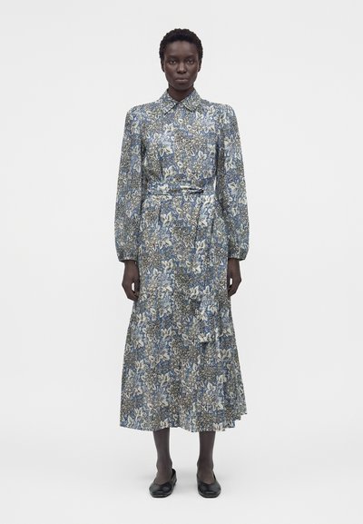 Femme debout de face, portant une robe florale à manches longues, longueur cheville, avec col et ceinture à la taille, associée à des chaussures plates noires.
