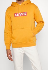 Žlutá mikina s kapucí vyrobená z bavlněné směsi, s předními kapsami ve stylu klokánky, žebrovanými manžetami a červenou nášivkou loga Levi's na hrudi.