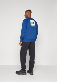 Blå sweatshirt med et hvidt logo på ryggen, parret med sorte sweatpants og sorte sportssko. Glat stof, afslappet pasform.