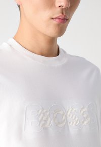 Weißes Baumwoll-T-Shirt mit einem gerippten Rundhalsausschnitt und einem geprägten Design, das das Wort "BOSS" in hellbeige zeigt.
