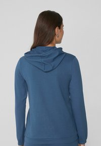 Mujer con pelo largo castaño que lleva una sudadera azul con capucha, vista de espaldas contra un fondo sencillo.
