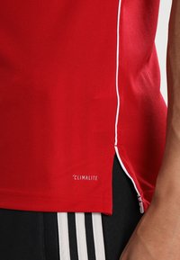 Rotes Sport-T-Shirt aus strukturiertem Stoff, mit weißen Paspeln an den Seiten und einem 'CLIMALITE'-Label nahe dem Saum.