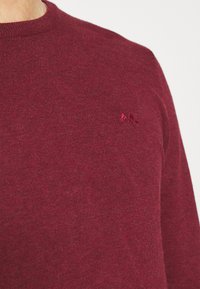 Burgunderfarbener Pullover mit Rundhalsausschnitt, aus weichem Material gefertigt. Mit einem kleinen gestickten Logo auf der Brust und einer glatten, gleichmäßigen Textur.