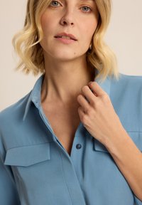 Jasnoniebieska koszula z długim rękawem, z kołnierzykiem, dwiema kieszeniami na piersi i miękką teksturą materiału. Model dotyka kołnierzyka na neutralnym tle.