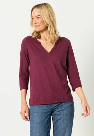 Woman with shoulder-length blonde hair wearing a maroon long-sleeve V-neck shirt and blue jeans, looking to her left.
