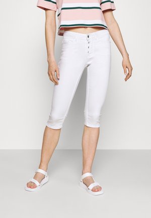 Personne portant un pantalon skinny blanc court avec braguette à boutons, un haut court rose à rayures vertes, et des sandales blanches à bout ouvert.