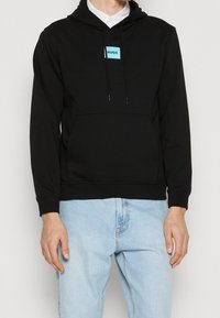 Sudadera con capucha negra con bolsillo tipo canguro, que presenta un parche rectangular azul con el logo "HUGO." Diseñada con capucha ajustable mediante cordones y puños acanalados.