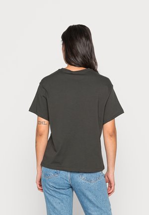 Frau mit langen dunklen Haaren, die ein schlichtes schwarzes T-Shirt mit kurzen Ärmeln und hellblaue Jeans trägt, steht mit dem Rücken zur Kamera.