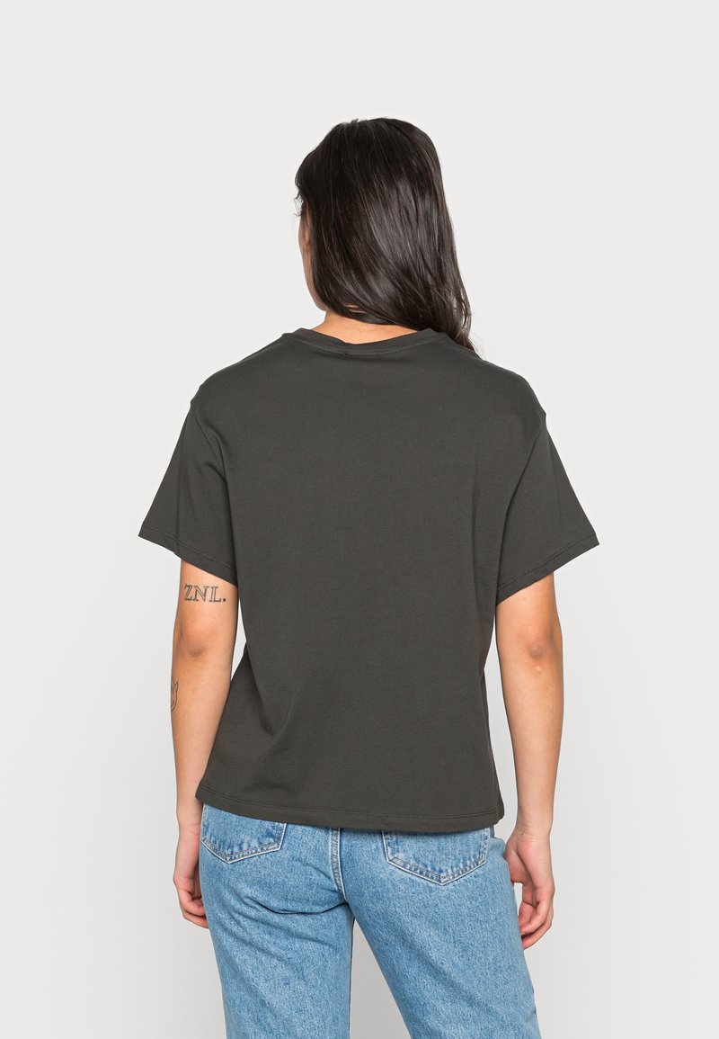 Femme aux longs cheveux foncés portant un t-shirt noir uni à manches courtes et un jean bleu clair, debout de dos face à l'appareil photo.