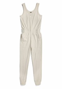 Ärmlös beige jumpsuit med elastisk midja, sidofickor, ribbade ankle cuffs och liten vit logotyp på bröstet.