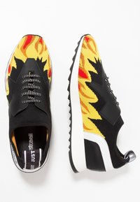 Zapatillas negras con un diseño de llamas amarillas y rojas, textura tejida, acentos negros en contraste y una suela de goma blanca.