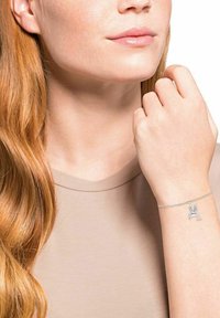 Mujer con cabello largo y rojo que lleva una blusa beige y una pulsera de plata con un charm de ave voladora en su muñeca.