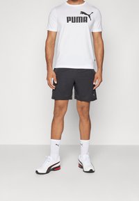 Hombre vistiendo una camiseta blanca de Puma, pantalones cortos negros, calcetines blancos de Puma y zapatillas blancas, de pie frente a un fondo blanco liso.