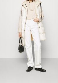 Kvinna som bär en krämfärgad tröja, vit vadderad lång väst, vita byxor, svarta skor och håller i en liten svart handväska med kort rem.