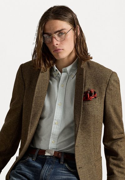 Blazer en chevrons marron sur une chemise à rayures bleu clair, featuring un pochette de poche à motifs rouges et un jean en denim foncé, accentué par une ceinture marron.