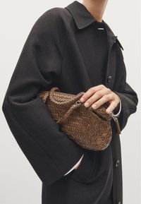 Braune geflochtene Handtasche, die in einer Hand gehalten wird, mit einer runden Form, Reißverschluss und dekorativem Quast, kombiniert mit einem schwarzen Mantel.