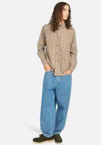 Braun kariertes Hemd mit langen Ärmeln, kombiniert mit locker sitzenden hellblauen Jeans und braunen Wildlederschuhen. Minimale Accessoires.