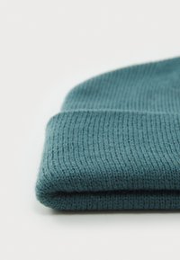 Gorro de malha teal com textura canelada, dobra na bainha e material macio. A superfície é suave, com uma cor uniforme e profundidade subtil.