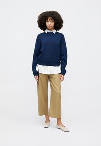 Pull en tricot bleu marine sur une chemise blanche à col, associé à un pantalon large beige clair et des ballerines beige pâle. Textures lisses, style décontracté.