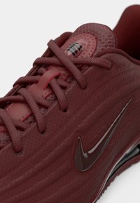 Tuvumā redzams tumši sarkans sporta apavu modelis ar strukturētu audumu, šņorēm un spīdīgu Nike zvīņas logotipu sānos.
