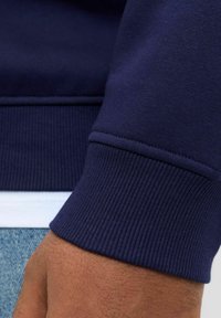 Primer plano de la muñeca de una persona que lleva un sudadera azul marino con puños de canalé, una camiseta blanca y jeans azules.