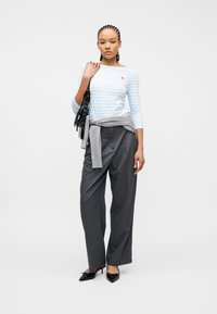 Γυναίκα με πλεξούδες που φοράει ανοιχτό γαλάζιο ριγωτό τοπ, γκρι φαρδιά παντελόνια, μαύρες γόβες, γκρι πουλόβερ δεμένο στη μέση και κρατά μαύρη τσάντα.