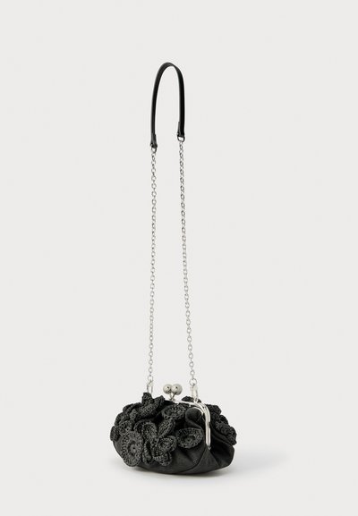 Μαύρο υφασμάτινο πορτοφόλι με λεπτομέρειες από πλεκτό λουλούδι, ασημένιο κούμπωμα και μακρύ ασημένιο λουράκι με μαύρη δερμάτινη επένδυση στον ώμο.