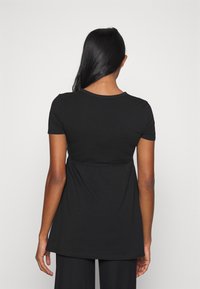 Mujer con largo cabello negro vestida con una blusa corta y holgada negra y pantalones negros, de espaldas contra un fondo liso claro.