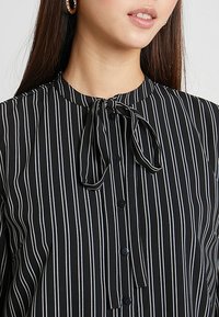 Blusa a righe bianche e nere con chiusura a bottoni, colletto con dettaglio a fiocco e texture di tessuto liscia.