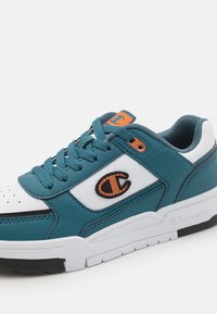 Sneaker teal e bianco con punta perforata, dettaglio arancione del logo, lacci grigi e superficie texturizzata. Presenta una robusta suola bianca e accenti neri.