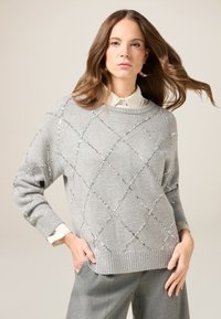 Grauer gestrickter Pullover mit Rautenmuster, verziert mit silbernen Pailletten, ausgestattet mit einem gerippten Saum und weiten Ärmeln über einem weißen Hemd mit Kragen.