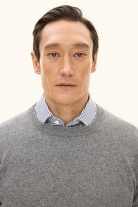 Grauer Pullover aus einem weichen, strukturierten Stoff, über einem gestreiften weißen und blauen Hemd mit Kragen getragen. Die Umgebung ist neutral und minimalistisch.