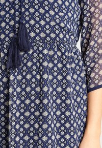 Marinblått klänning i tyg med ett mönster som har vita blommotiv, elastisk midja och dekorativa mörkblå fransar.