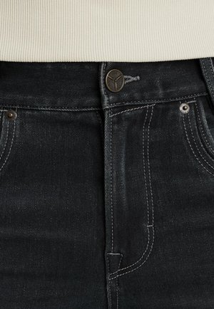 Detailný záber na pás čiernych denimových nohavíc s kovovým gombíkom, pútkami na opasok a bielym rebrovaným sveterom zasunutým dovnútra.