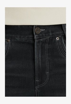Dettaglio ravvicinato della vita di jeans in denim nero con bottone in metallo, passanti per cintura e maglione bianco a costine infilato dentro.