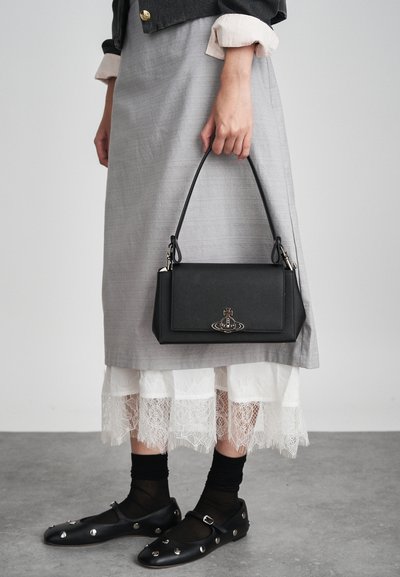 Melna struktūrēta rokassoma ar sudraba logo, turēta rokā. Valkāta ar pelēku svārku un baltu mežģīņu apakšslāni. Strukturēti melni apavi.