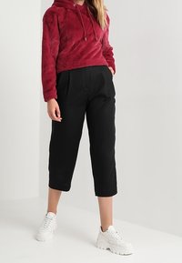 Felpa rossa in peluche con cappuccio e lacci, abbinata a pantaloni neri corti e sneakers bianche chunky. Texture liscia e vestibilità ampia in tutto.