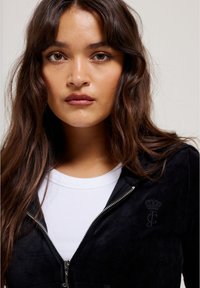 Black zip-up hoodie with plush texture and embroidered logo; layered over a white ribbed shirt. Wavy hair frames the face.