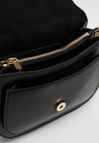 Mustassa nahkaisessa crossbody-laukussa on kultainen vetoketju ja napitus, ja siinä on useita lokeroita sekä mokkanahkainen sisustus.