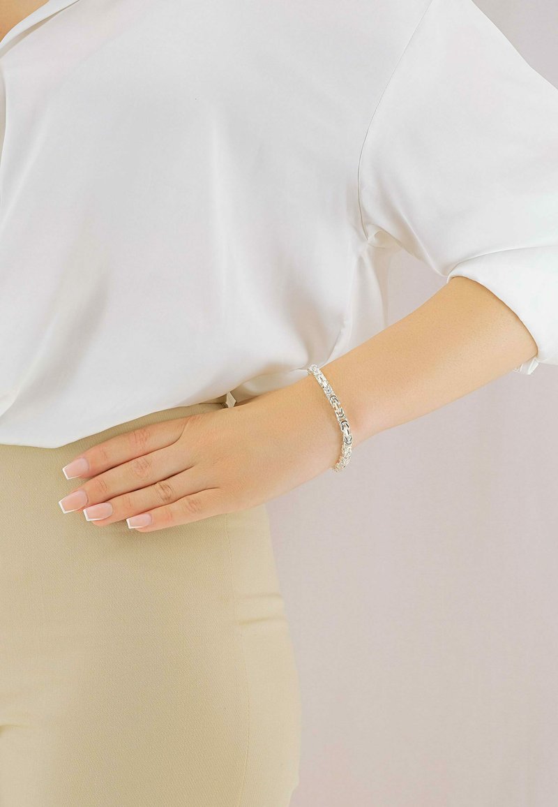 Silbernes Armband mit filigranen floralen Mustern, getragen an einer Hand, die auf einem beigen Gürtel ruht, vor einem hellen Hintergrund.