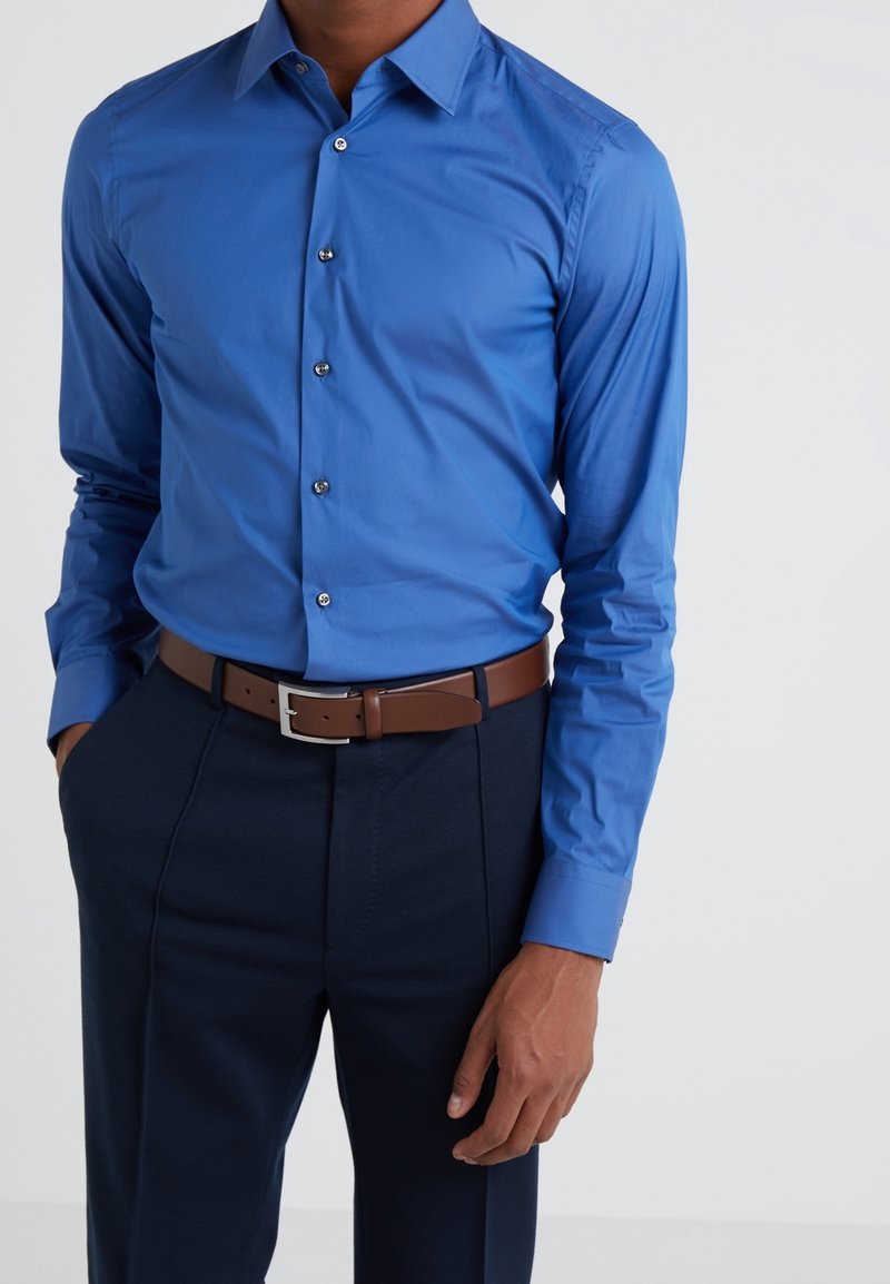Homme portant une chemise ajustée bleu vif à manches longues boutonnée, rentrée dans un pantalon foncé avec une ceinture marron, debout devant un fond uni.