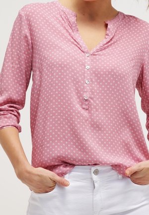 Femme portant un chemisier rose à manches longues avec de petits motifs blancs et un pantalon blanc, mains dans les poches.