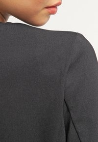 Zbliżenie dolnej części twarzy i ramienia osoby w czarnej teksturowanej odzieży na jednolitym jasnym tle.