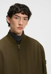 Olive green bomber jacket with a ribbed collar, zip closure, and side pockets. The fabric appears smooth with a slight sheen.